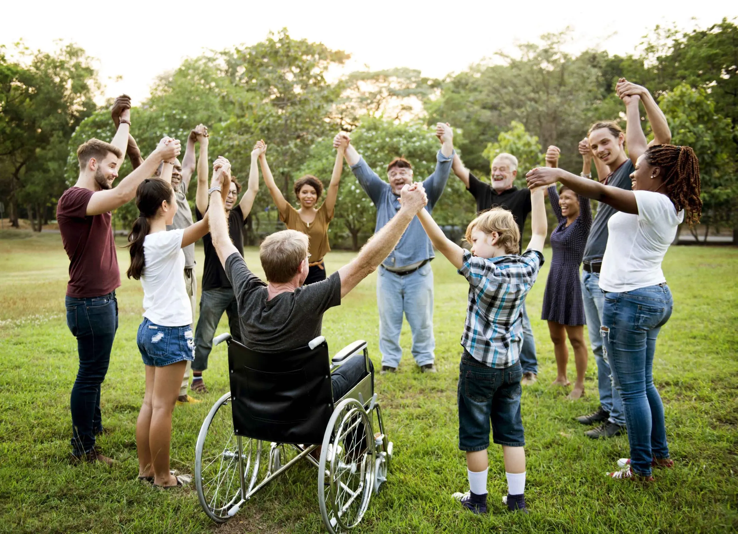 A mix of smiling adults and children stand in a circle in a park holding eachothers hands in the air. One man in the foreground is in a wheelchair